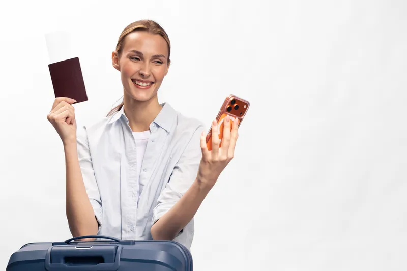 Happy woman holding passport and smartphone takes selfie with suitcase in front of white background. Concept of travel, vacation, preparation, and modern lifestyle with technology.