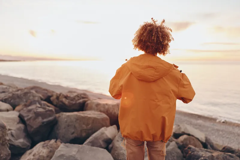 Woman lifestyle, hooded jacket, curly hair, standing on rocky beach by ocean at sunset, back view, calm walk, warm evening light, travel mood