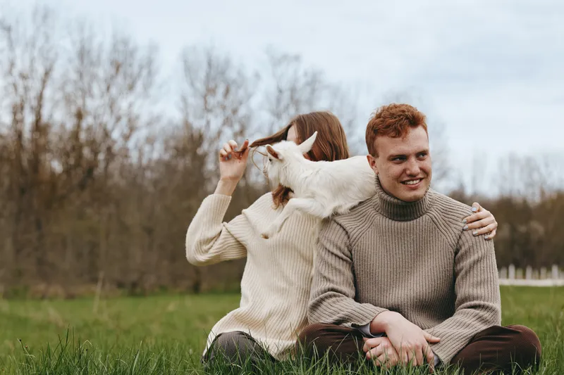 Cheerful couple outdoors with a playful goat kid nearby, relaxed moment on grass, soft sweater textures, natural light, intimate connection, rustic setting, candid lifestyle scene