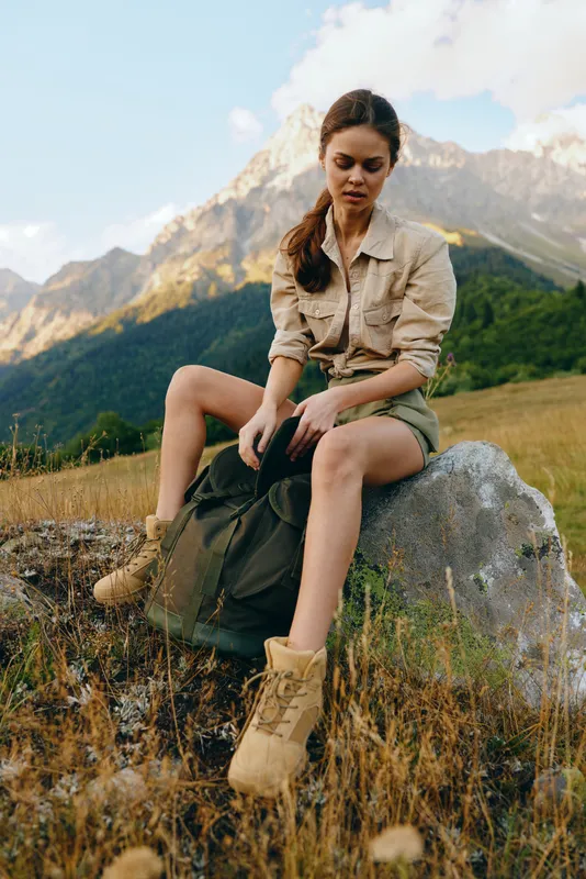 Casual woman outdoor explorer sits on a rock in the mountains wearing khaki shirt shorts boots backpack enjoying sunny day and vast horizon