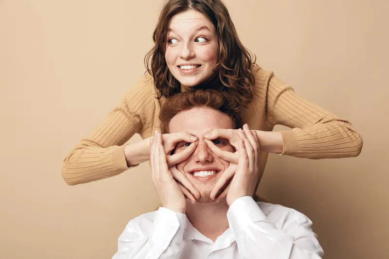 Happy young woman in brown sweater playfully covers eyes of smiling man in white shirt. Fun couple enjoying moment together on beige background. Concept of joy and relationship.