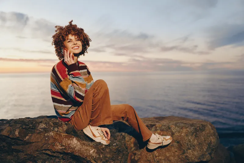 Woman lifestyle portrait at sunset on rocky coast, ocean horizon behind, curly hair, striped sweater, brown pants, thoughtful pose, hand near face, smartphone in lap, calm travel mood