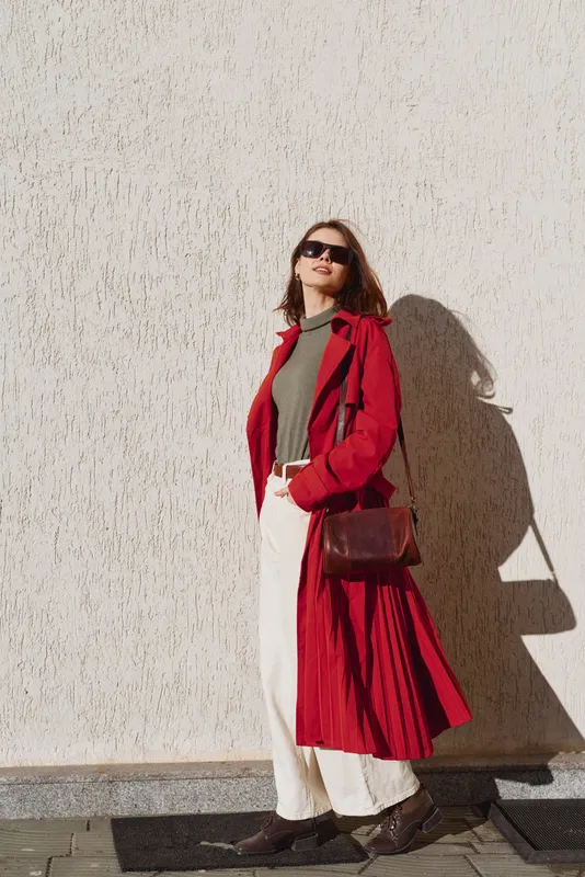A confident woman in a red trench coat and sunglasses stands against a textured wall, posing with a brown leather bag in bright sunlight.