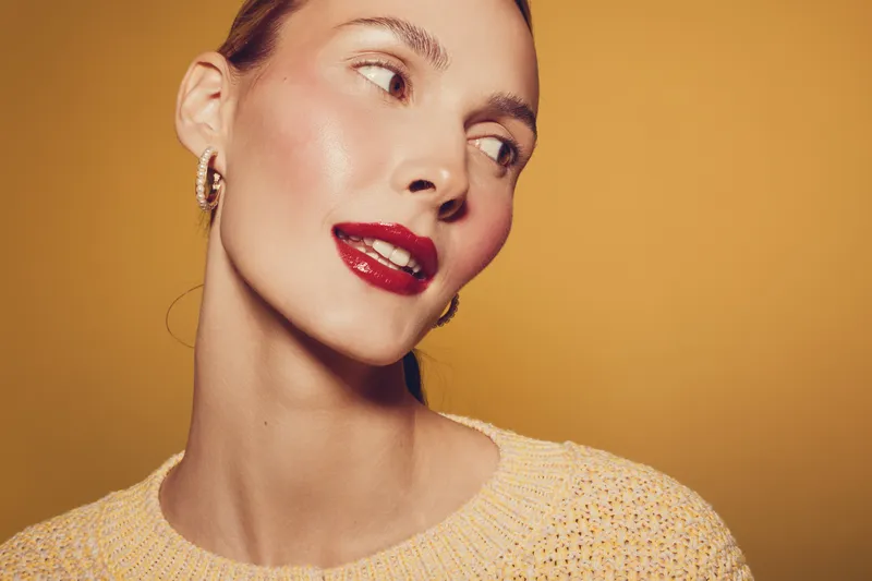 Close-up of a young woman with glossy red lips and smooth skin looking sideways on a warm yellow background. Concept of beauty, makeup, skincare, and fashion portrait.