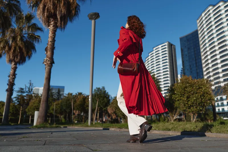Woman in a flowing red coat walks along a sunny city street with modern glass buildings, conveying bold style, movement and urban confidence.