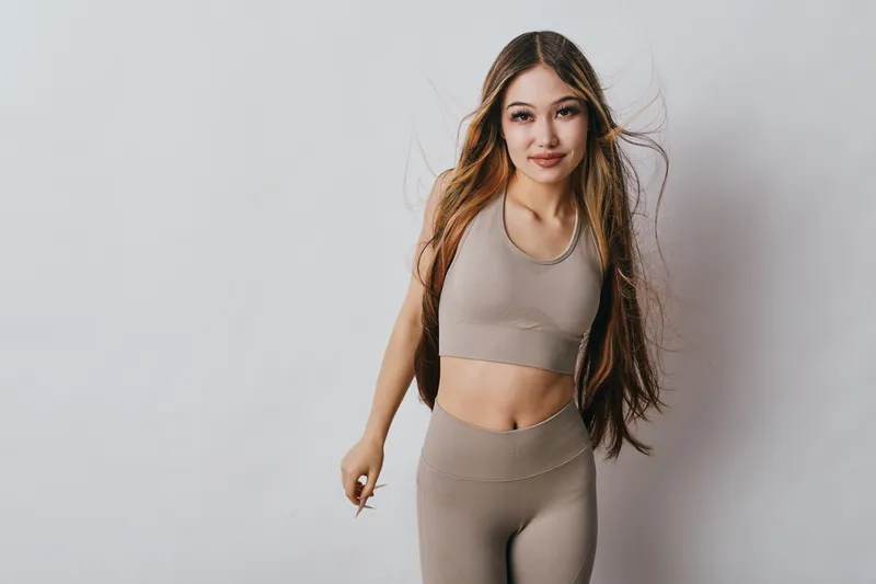 Young woman in beige sportswear posing with confident smile and long hair against plain background for fitness, health and active lifestyle concept image.
