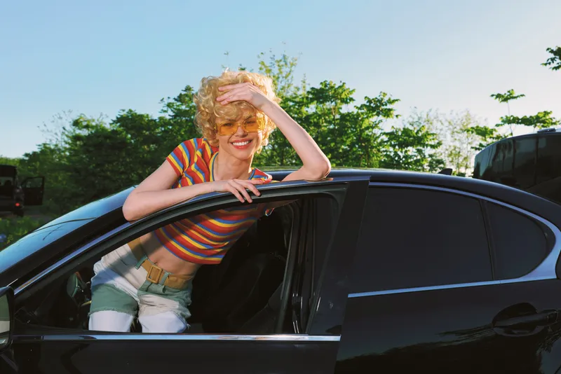 A cheerful woman leans out of a black car window, sunlit outdoors, wearing a striped shirt and denim shorts. Retro film color styling conveys a carefree lifestyle. Bright summer mood.