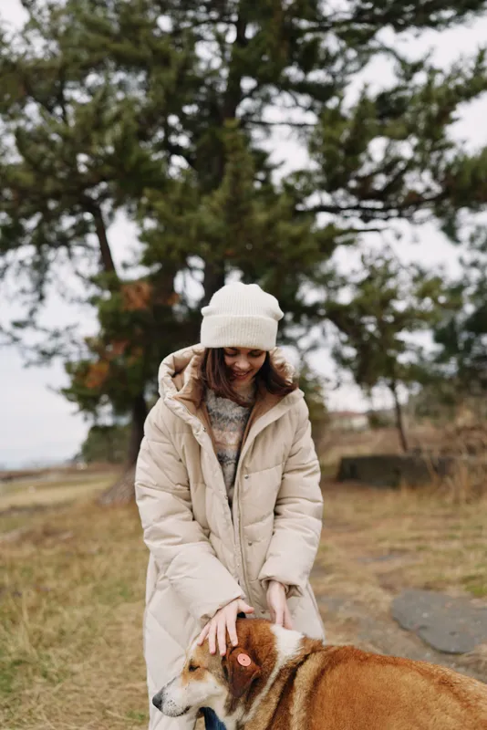 Woman in a long beige winter coat and knit hat outdoors with a large dog, strolling through a park with trees and grass, conveying a cozy, calm moment of companionship in cool weather.