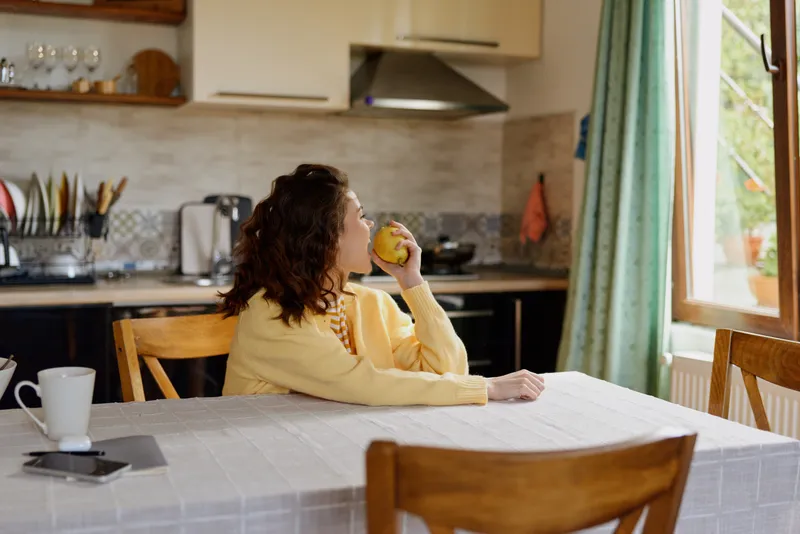 Curly haired woman in a yellow sweater sits at a dining table in a bright kitchen, sipping juice, capturing a warm casual home lifestyle moment with soft natural light.