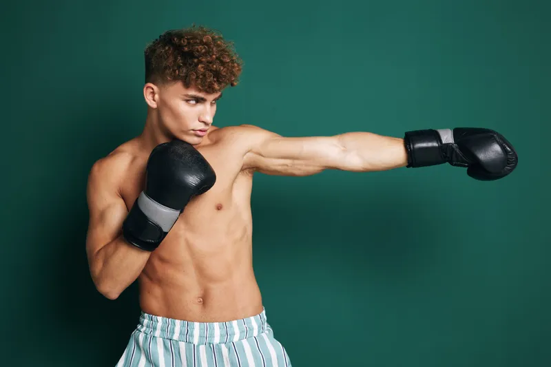 Young man with boxing gloves training and punching with bare torso in striped shorts on green background for fitness and sport workout.