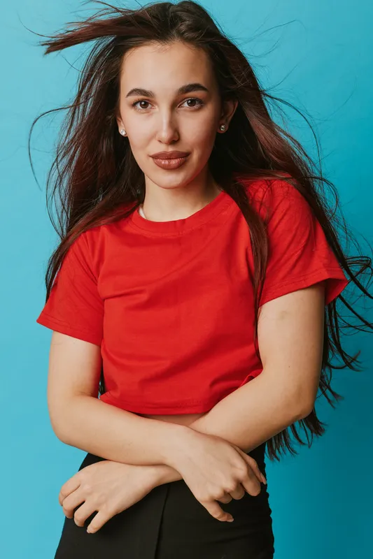 Young woman with long dark hair wearing red shirt posing with crossed arms and slight smile against blue background. Casual style, confident look and natural beauty in studio portrait.