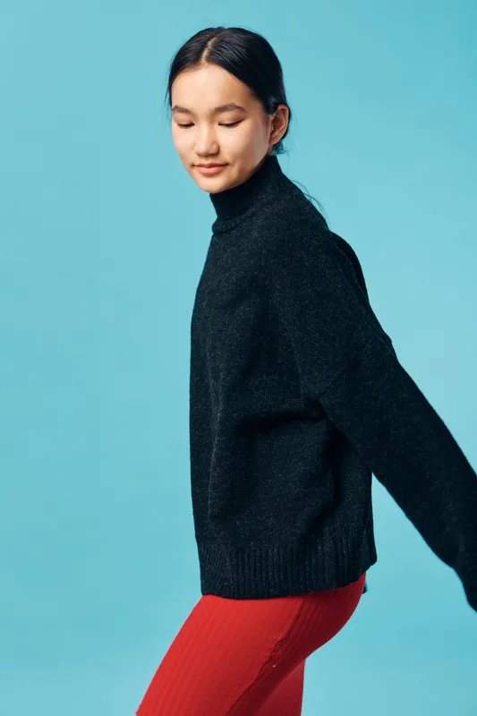 Young woman with dark hair in black sweater and red pants, posing with eyes closed against solid blue background, fashion portrait with casual style and calm expression.