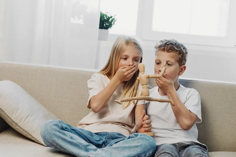 Two children, boy and girl, sitting on sofa playing with wooden puppet. They cover their mouths with hands, showing surprise and curiosity in bright living room with natural light.