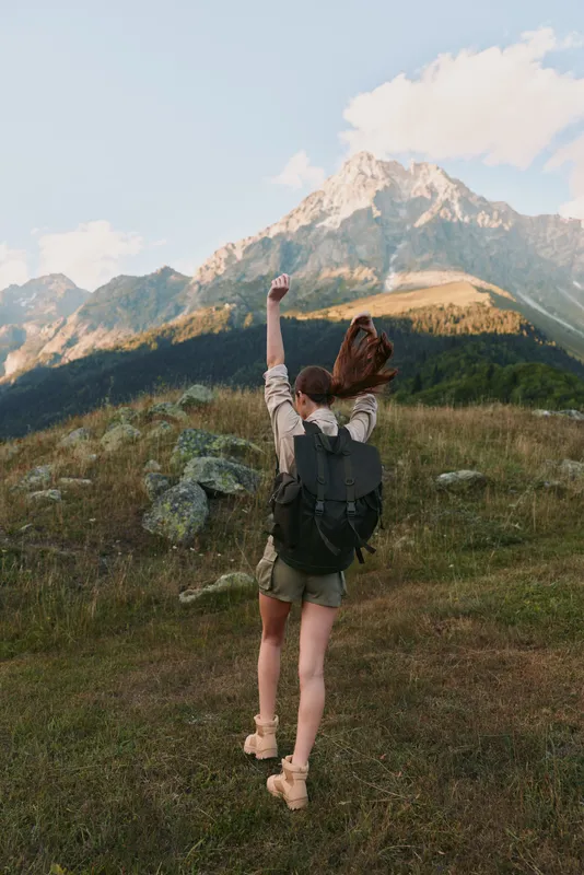 Young woman hiking outdoors with backpack celebrating success in mountain landscape, casual outfit, sunny day, natural environment, active lifestyle concept