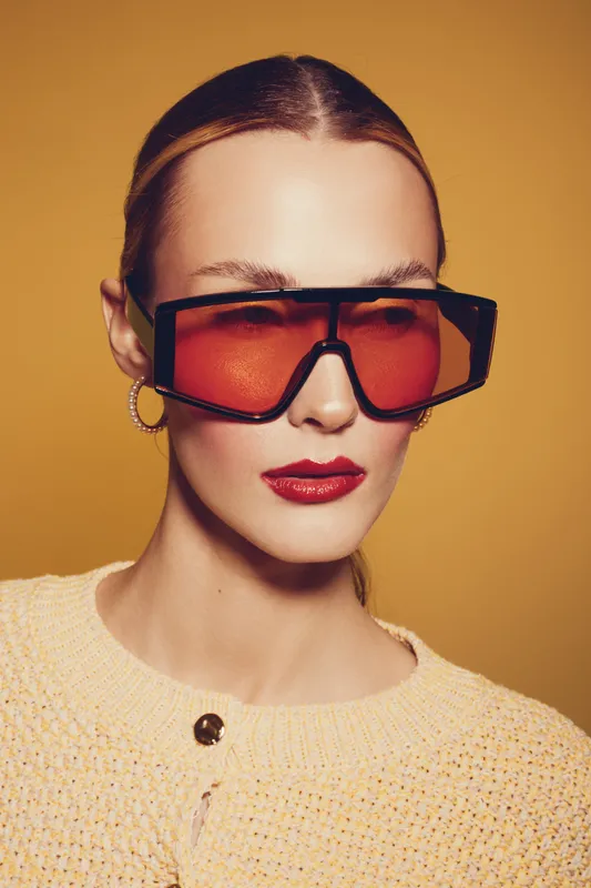 Stylish woman with slicked back hair wears oversized red sunglasses and hoop earrings. She poses confidently against a warm yellow background in a textured cream sweater.