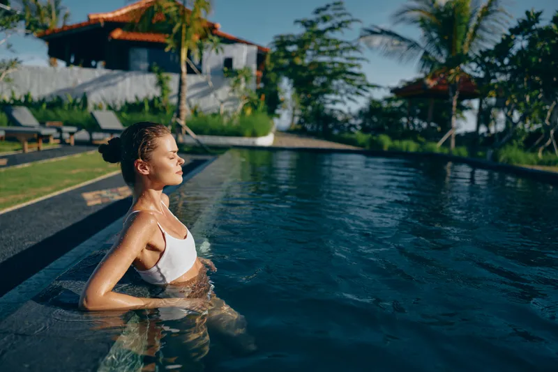 Sunlit pool moment shows a young woman in a white bikini standing in the water with eyes closed, exuding calm as reflections shimmer on the surface.