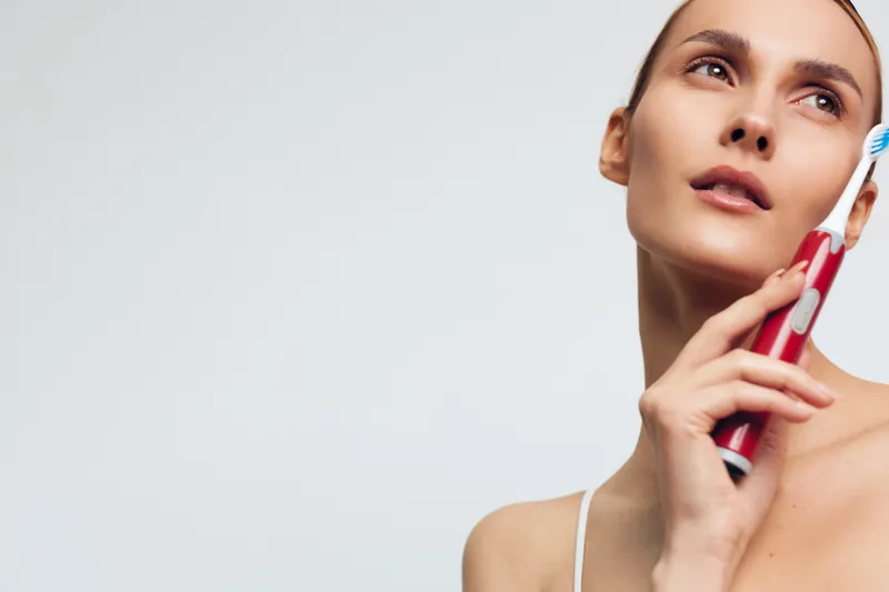A young woman holds an electric toothbrush near her face looking upwards with a neutral expression against a plain white background. Concept for dental care and personal hygiene.