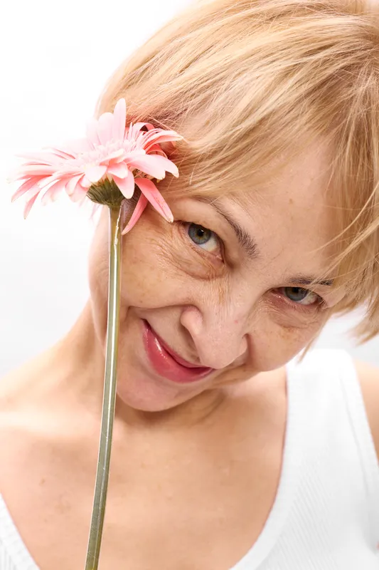 Woman closeup smiling, pink flower near face, blonde hair, indoor bright light, soft skin texture, gentle expression, white background, beauty portrait