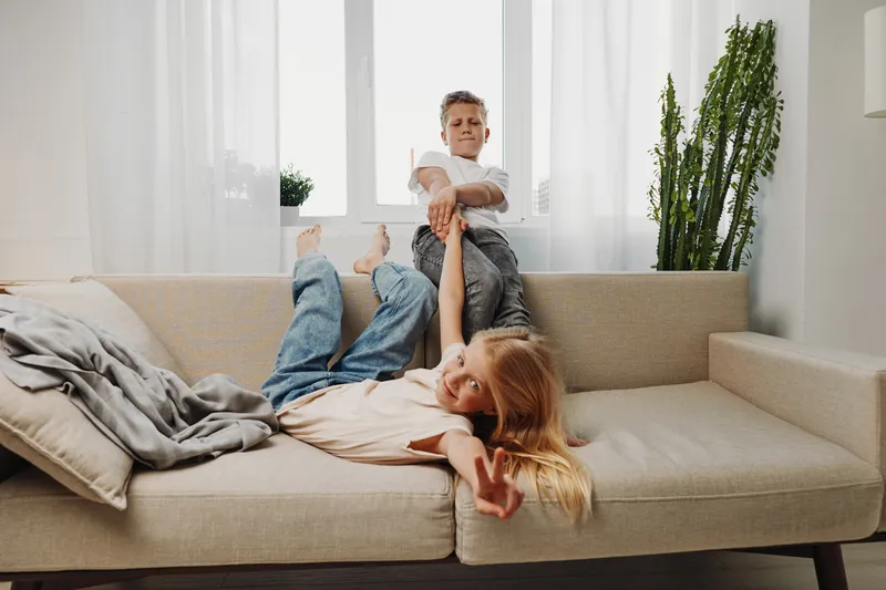 Playful siblings having fun on beige sofa in bright living room. Boy pulls girl’s arm as she lies smiling and making peace sign. Concept for family, childhood and joyful moments.