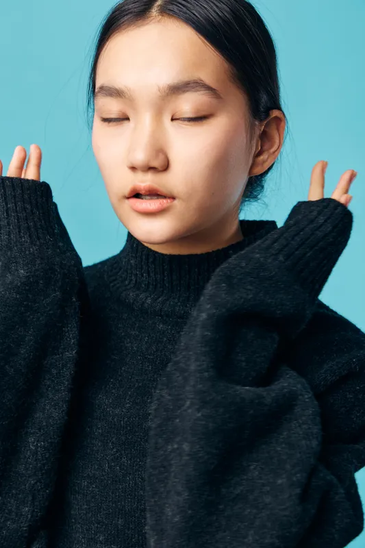 Young woman with closed eyes wearing black sweater posing with raised hands on blue background studio portrait, calm expression and dark hair tied back for fashion or relaxation.