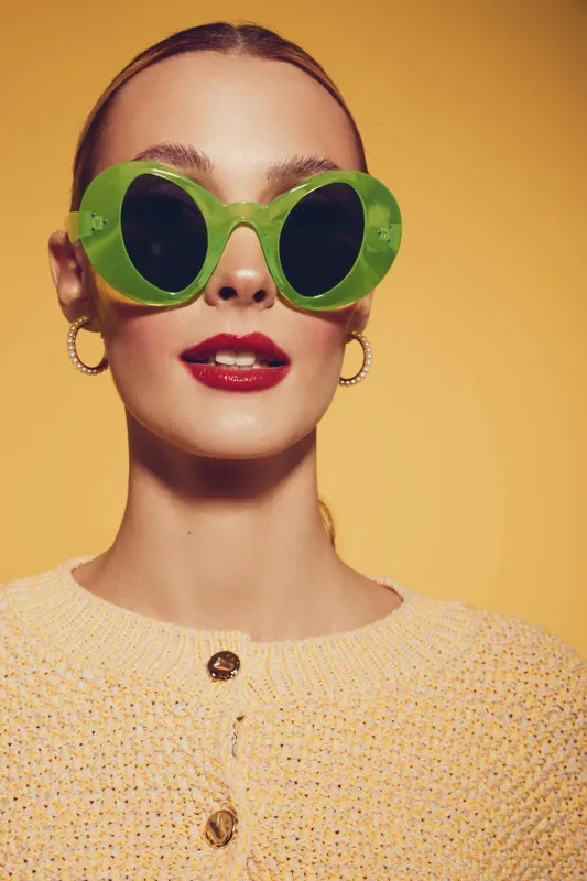 Young woman with green oversized sunglasses and red lipstick poses against yellow background. She wears a beige knitted sweater with buttons. Concept of fashion, style, and summer accessories.