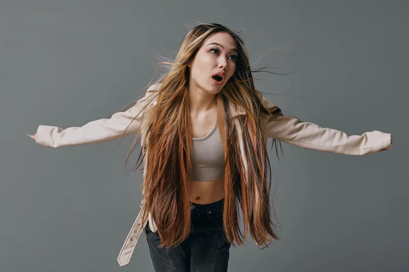 Young woman with long hair posing with open arms and expressive face on grey background for fashion, beauty and casual style portrait.