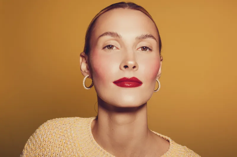 Close-up portrait of confident woman with red lipstick and hoop earrings wearing knitted sweater on yellow background. Concept of beauty, fashion, and self-expression.