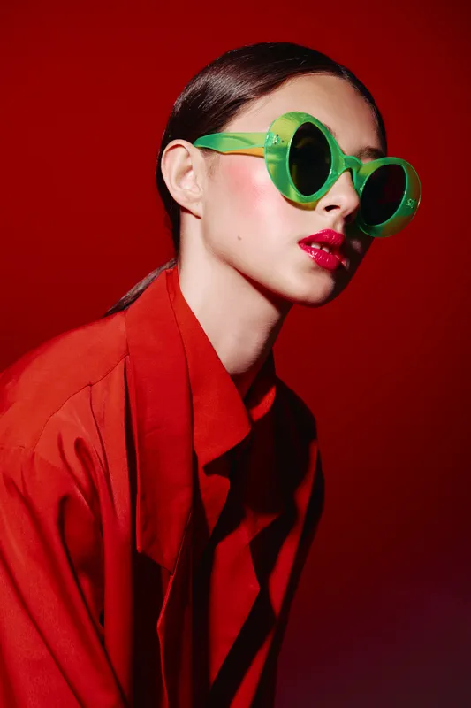 Fashion model with vibrant green sunglasses and glossy red lipstick poses against a red backdrop wearing an orange coat. Concept for beauty, creative fashion, and modern style.