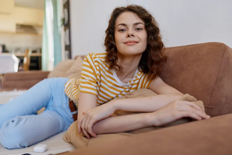 Cheerful woman relaxing at home on a sofa, striped shirt and casual vibe, soft natural light bathing a cozy living room, a candid lifestyle moment of comfort and calm relaxed