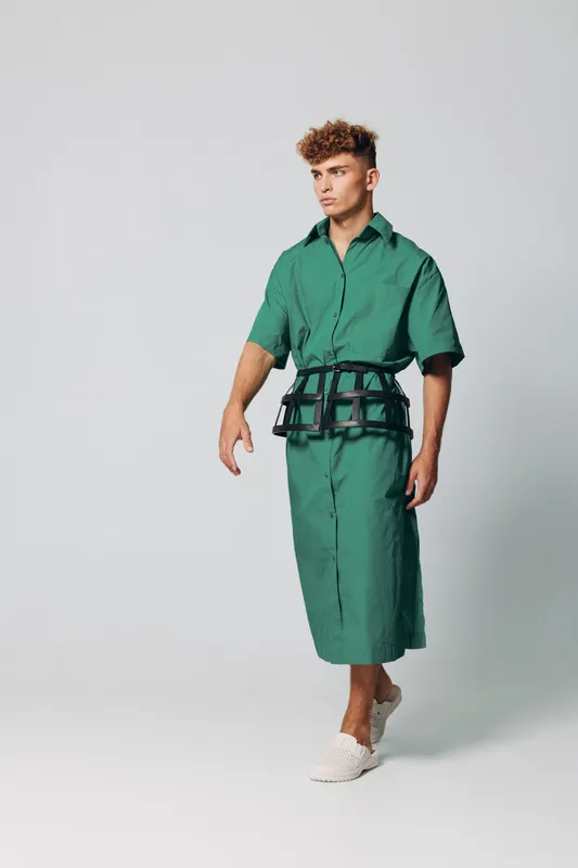Young man with curly hair wearing green shirt dress and black harness walking on white background. Fashion model posing in casual outfit with white sneakers in studio setting.