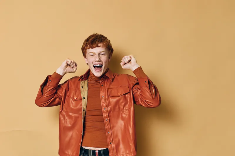 Young man with red hair and orange jacket stretching and yawning with closed eyes in front of beige background for morning routine and energy boost.