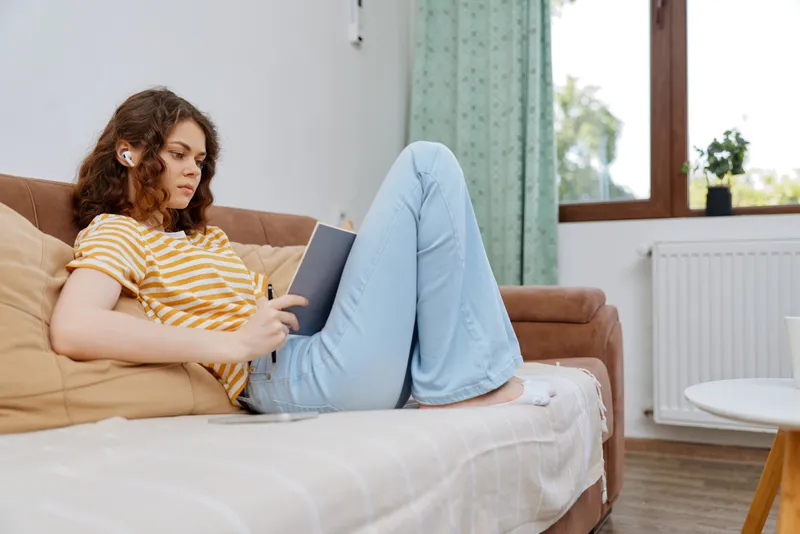 Casual woman sits on a sofa at home, reading on a tablet with a focused expression, natural daylight streaming through the window, cozy living room atmosphere.