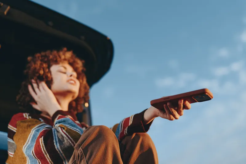 Woman with curly hair wearing a hat holding a smartphone outdoors under blue sky sunshine Lifestyle moment for modern communication and leisure Smiling portrait with warm sunlight and striped sweater