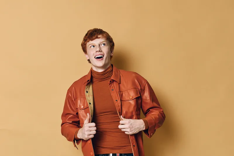 Young man with red hair wearing brown leather jacket and turtleneck stands against beige background looking up with smile and excitement in studio portrait.