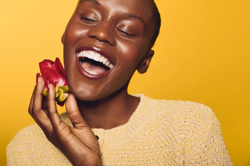 Happy African woman with dark skin laughing joyfully while holding a red flower near her face. Studio portrait on yellow background. Concept of natural beauty, wellness, and happiness.