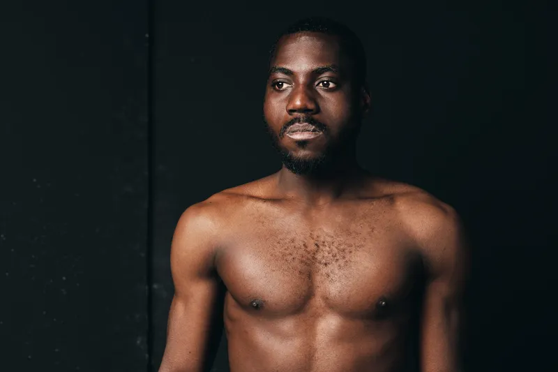 African man shirtless with muscular body and serious expression in creative conceptual visual style. Dark skin, portrait with studio lighting and black background for artistic photography.