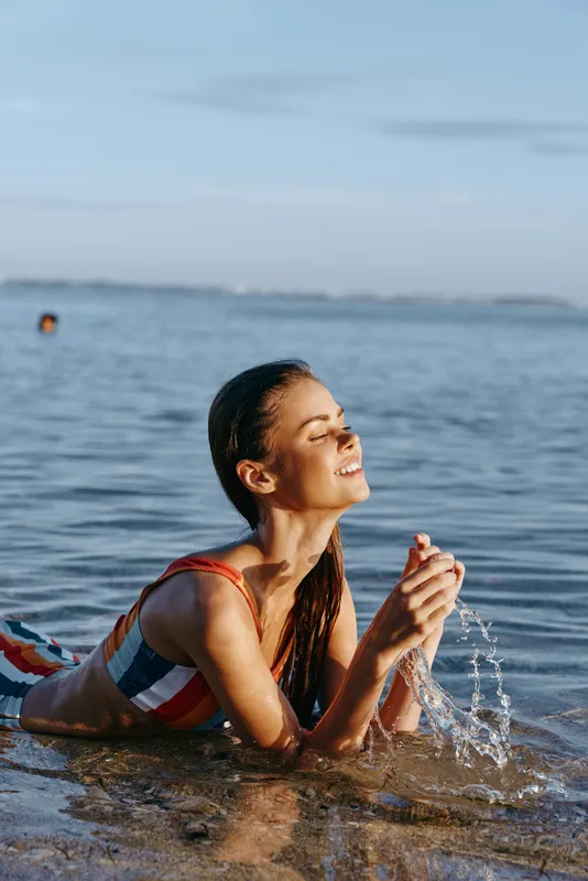 Woman swimming in ocean summer sunlight, smiling and laughing in shallow water, splashing droplets around her, wearing colorful swimsuit, joyful vacation mood by seaside horizon, fresh breeze