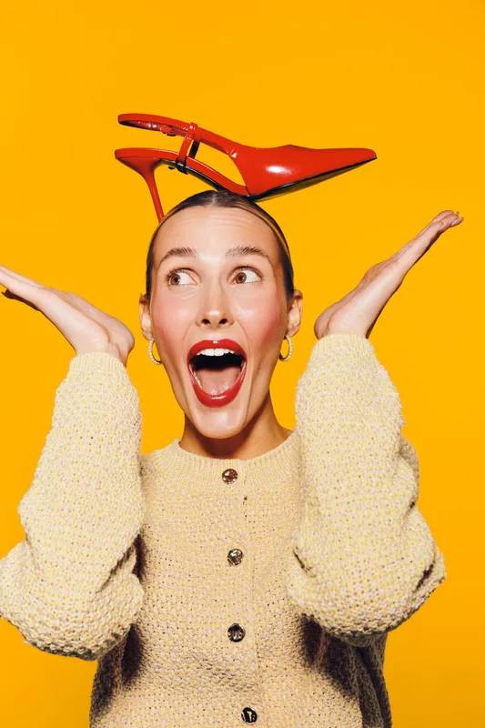 Young woman with red high heel shoe on head looks surprised and excited. She wears a beige sweater and gold earrings on bright yellow background. Concept of fashion, fun, and creativity.