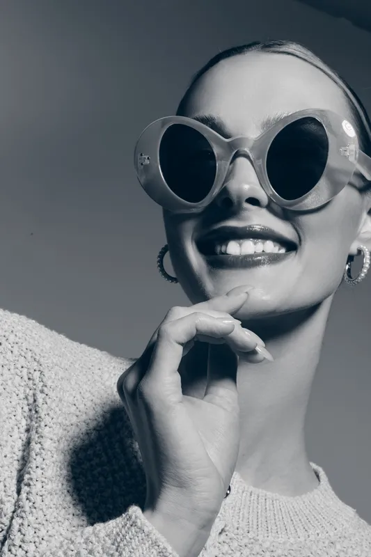 Smiling white woman with light skin wears large round sunglasses and wool sweater, posing with hand near face.