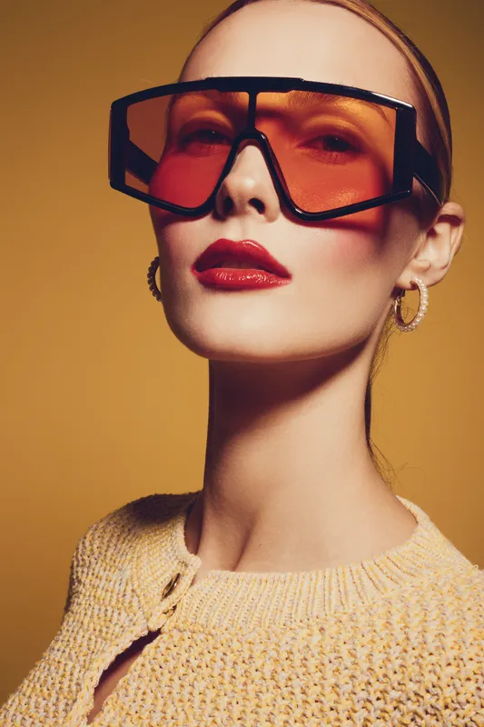 Stylish woman with oversized orange sunglasses and red lipstick poses confidently against a warm yellow background. Fashion concept with bold accessories and makeup.