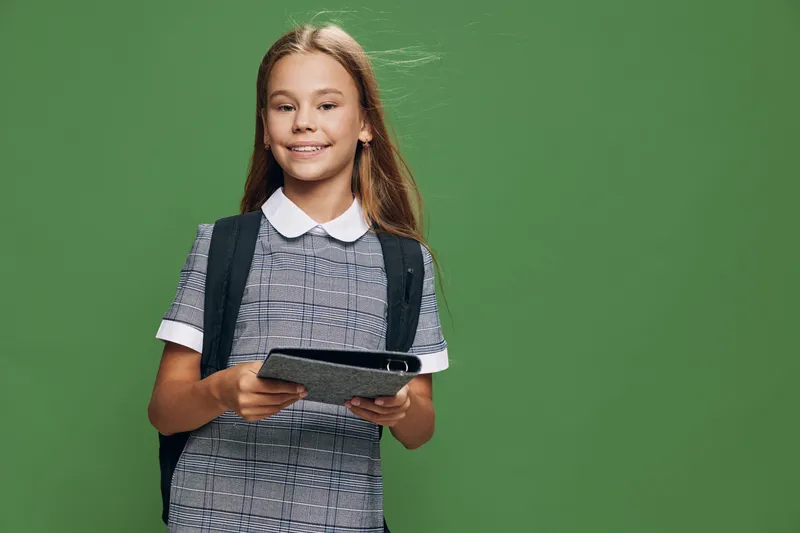 Schoolgirl with backpack holding digital tablet smiling and standing against green background in studio. Girl in uniform with long hair posing looking at camera with happy expression.