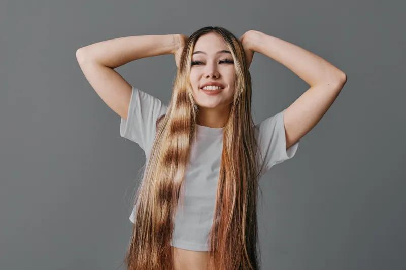 Young woman with long straight hair wearing casual white top posing with hands behind head smiling against gray background in studio. Female model beauty expression and confidence.