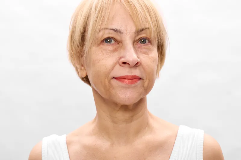 Mature woman portrait with blonde short hair, studio white background, calm neutral facial expression, soft light, skincare concept, close up shoulders