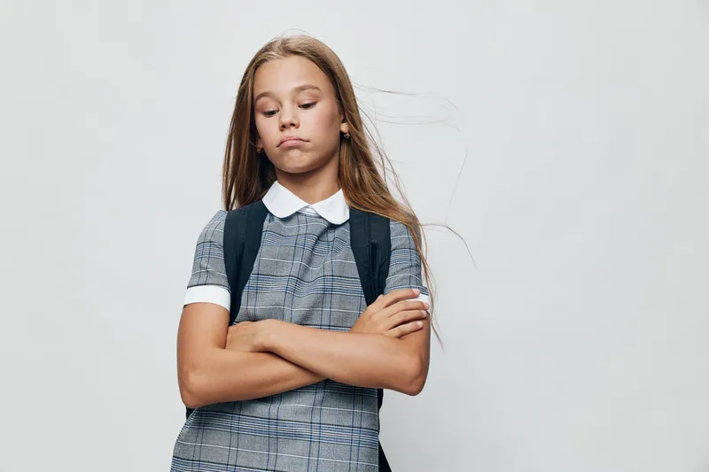 Girl student with backpack and crossed arms standing with closed eyes and serious expression against white background in school uniform for education concept.
