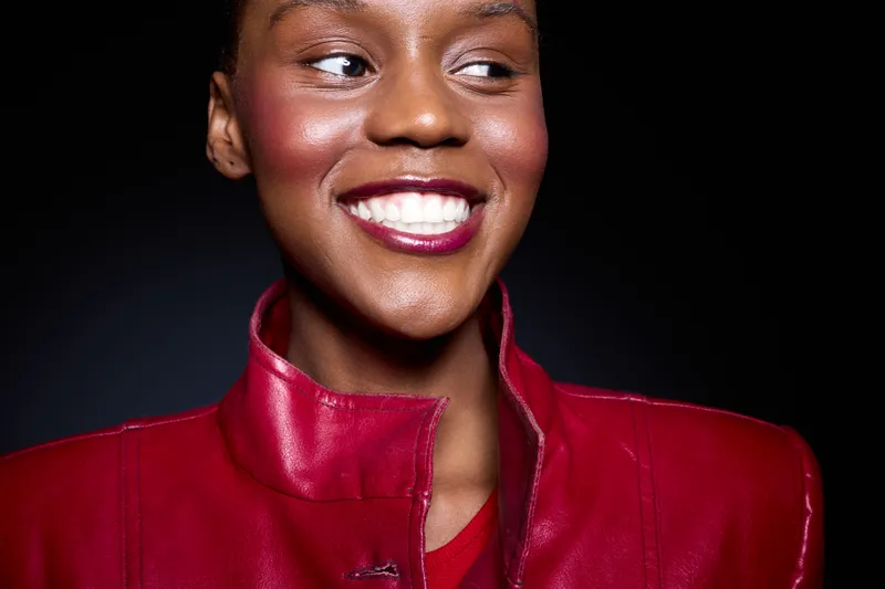 Close-up portrait of a smiling young black woman with dark skin wearing a red leather jacket against a black background. Concept of happiness, fashion, and beauty in studio lighting.