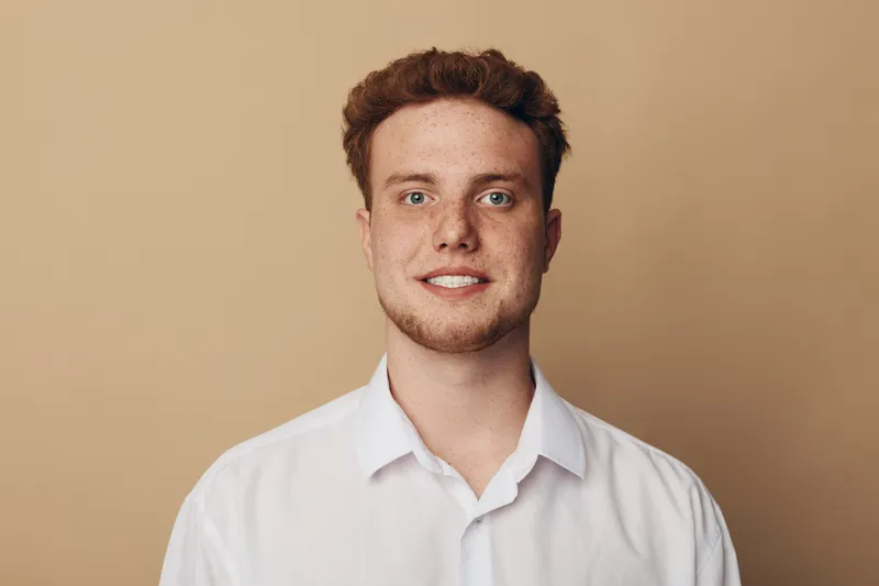 Young man with red hair and freckles smiles wearing white shirt against beige background. Portrait of confident male model with casual style and natural expression in studio lighting.