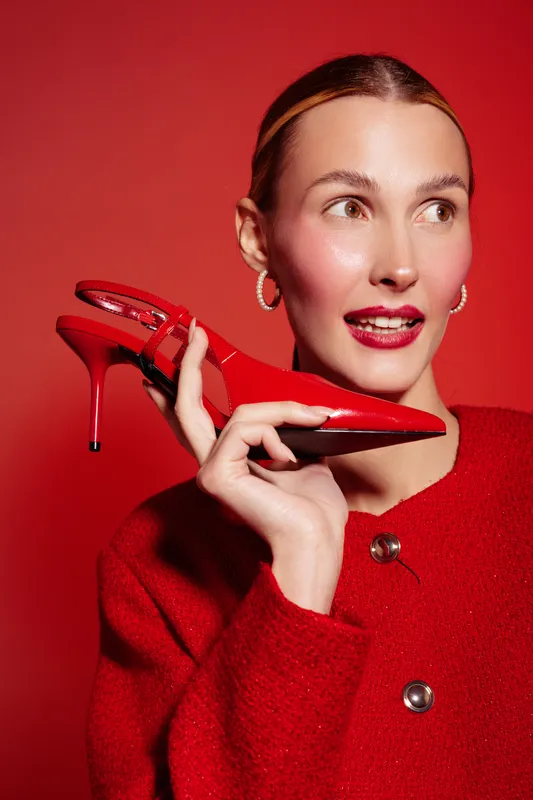Stylish woman in red sweater holds red high heel shoe near face, looking aside. Bright red background and matching lipstick create bold fashion and beauty concept with confident mood.