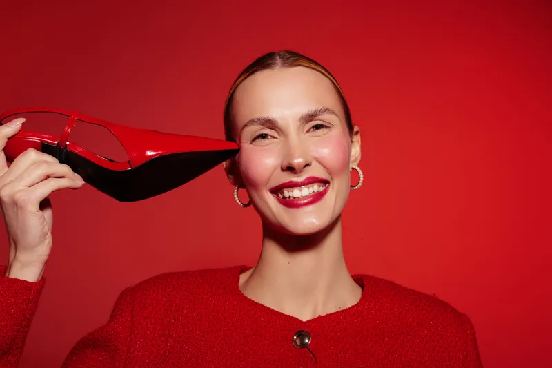 Smiling woman in red sweater holds red high heel shoe near face on red background. Concept for fashion, style, beauty, and confident female model with bright makeup and accessories.