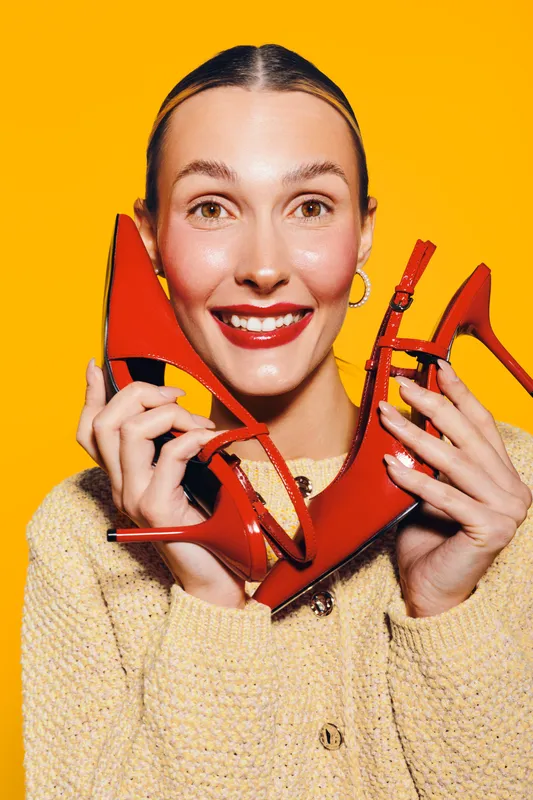 Smiling woman holds red high heel shoes near face on yellow background. Fashion concept with bright colors, beauty, and stylish footwear for trendy outfit and happy mood.