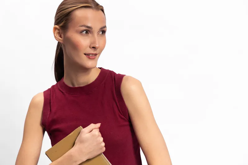 Young woman in a sleeveless red top holds a book close to her chest, looking sideways with a slight smile. Isolated on white background, concept for education, reading, and thoughtful mood.